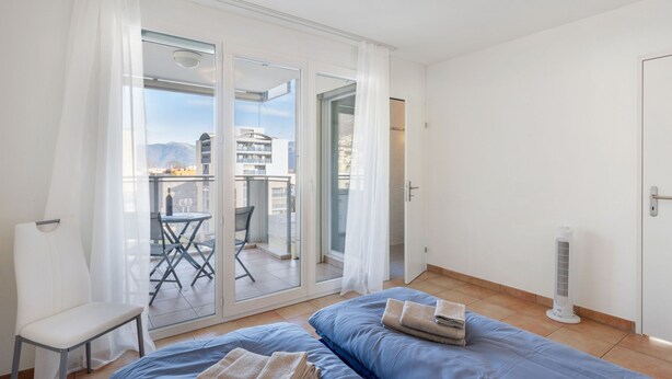 Une chambre à coucher comprenant un lit, une chaise, une table et un balcon offrant une vue sur les bâtiments et les montagnes.