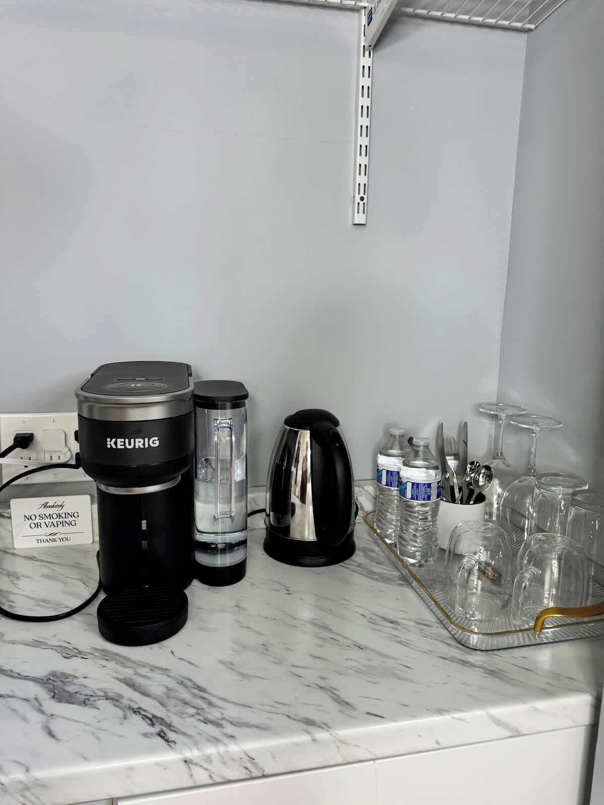 A kitchen counter with a Keurig coffee maker, a water dispenser, a kettle, and a tray with glasses and utensils.