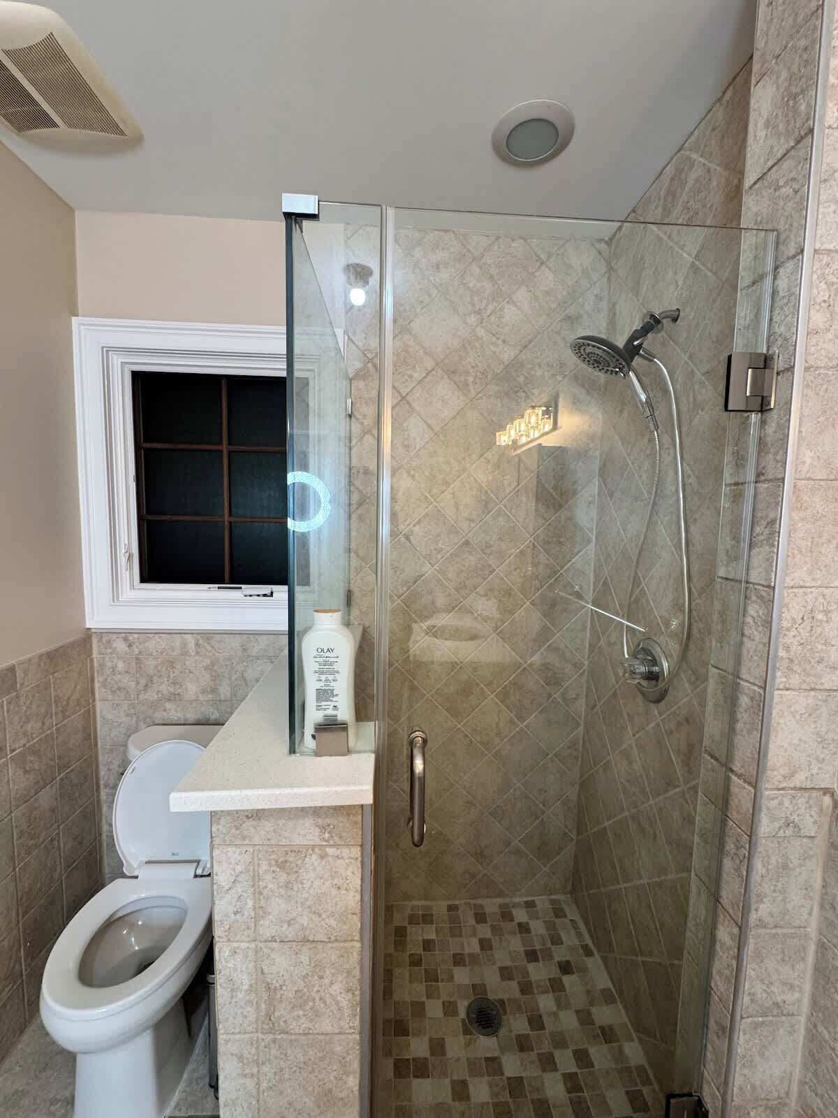 A bathroom with a glass-enclosed shower, a toilet, and a window above the sink.