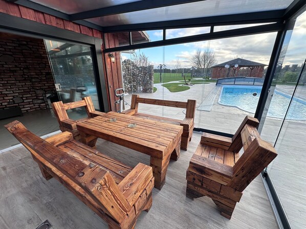 Et moderne udendørs spiseområde med et træbord, bænke, store vinduer og udsigt til en swimmingpool.