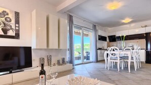A modern living room with a flat-screen TV, a dining area with white chairs, and a kitchen with dark cabinets.