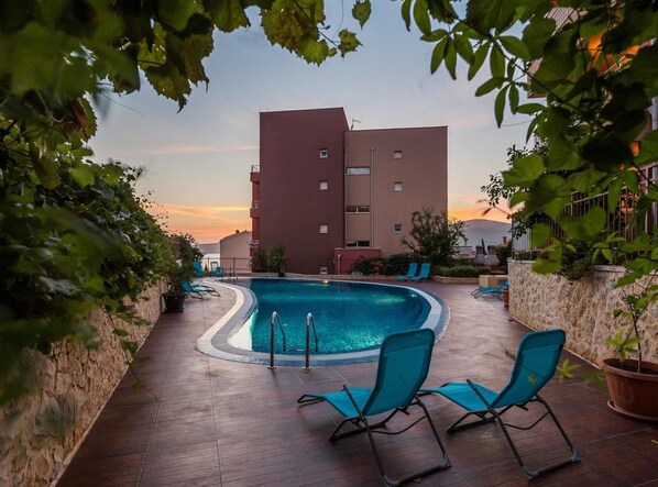 A swimming pool with lounge chairs, surrounded by a stone wall and vegetation, with a multi-story building in the background. - Luxury Apartment Okrug Gornji With Sea View and Pool (Okrug)