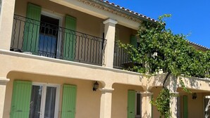 Un edificio de dos plantas con un balcón, contraventanas verdes y una mesa con sillas afuera.