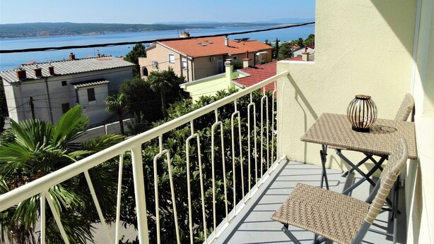 A balcony with a wicker chair and a table, overlooking a coastal town with red-roofed buildings and a clear blue sea. - Crikvenica Tranquil and Stylish Apartments With Sea View (Crikvenica)
