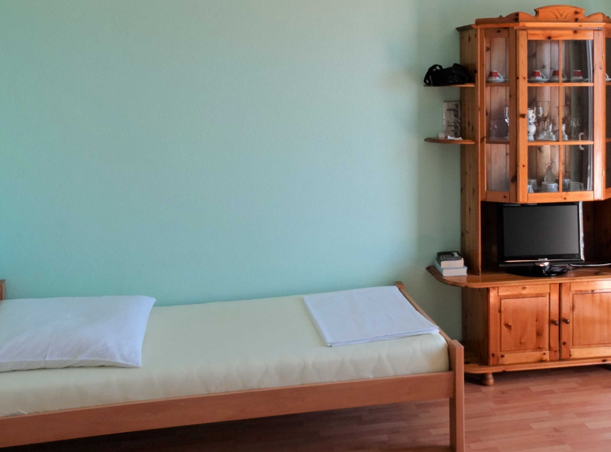 A single bed with a white sheet and pillow, a wooden cabinet with glass doors, and a television on a wooden stand.
