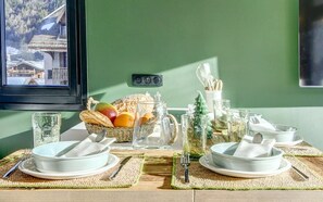 Een eettafel gedekt met borden, kommen, bestek, een broodmand, fruit en glazen, tegen een groene wand met uitzicht op een raam.