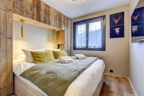 A bedroom with a wooden headboard, a large window with curtains, a bed with white linens and green pillows, and wall art depicting deer.