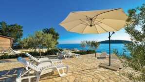A stone patio with white lounge chairs and a large beige umbrella, overlooking a clear blue sea and a distant island.