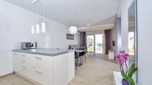 A modern kitchen with a central island, white cabinetry, and a microwave. There is a dining area with a table and chairs, and a view of the outdoors through a glass door.