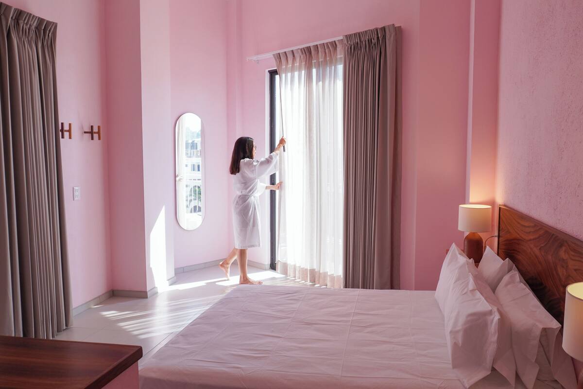 Quarto de hotel com cama grande, cabeceira de madeira, luminária de cabeceira e janela com cortinas.
