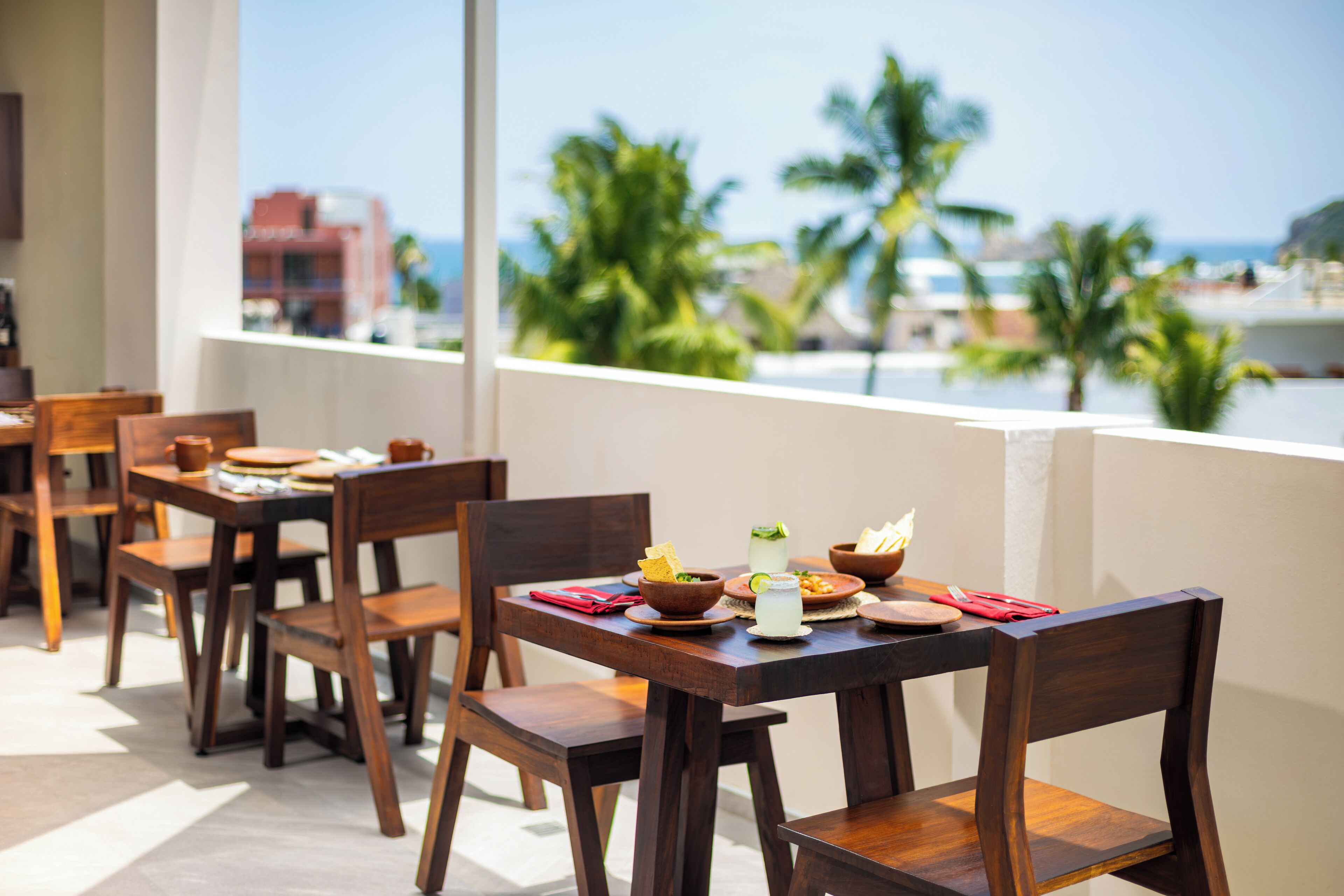 A dining area with wooden tables and chairs, a view of palm trees and the sea, and a building in the background.