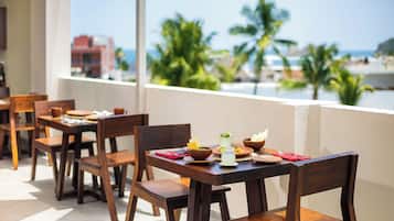A dining area with wooden tables and chairs, a view of palm trees and the sea, and a building in the background.