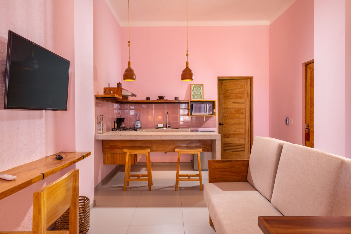 A compact living space with a kitchenette, a flat-screen TV mounted on the wall, wooden furniture, and a sofa.