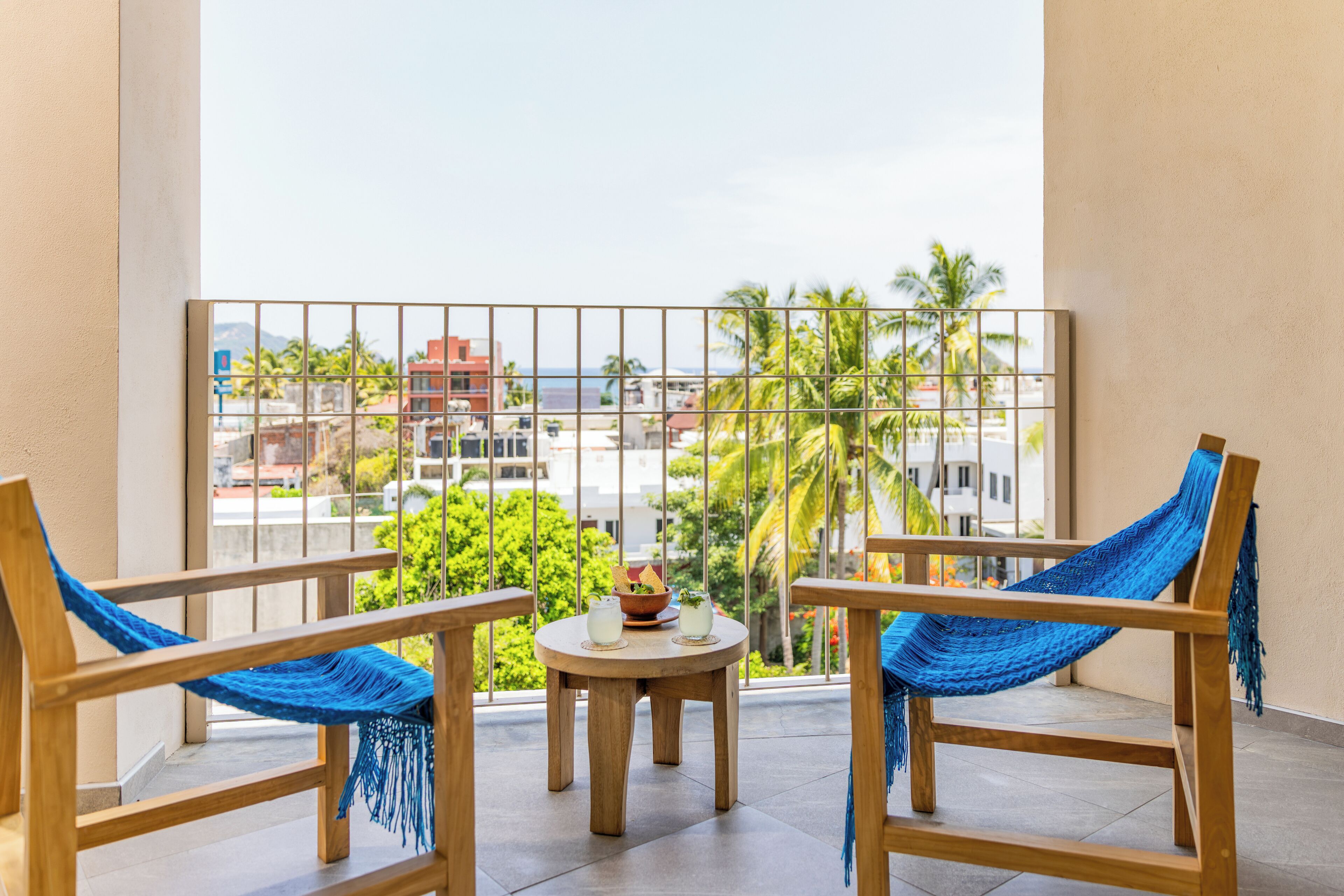 Un balcón con dos sillas de madera y una mesa redonda pequeña, con vista a palmeras y edificios.