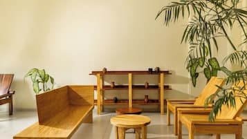Una habitación con un banco de madera, una mesa redonda pequeña y una silla. Hay una estantería con objetos decorativos y una planta grande.