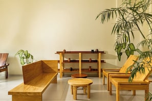Una habitación con un banco de madera, una mesa redonda pequeña y una silla. Hay una estantería con objetos decorativos y una planta grande.