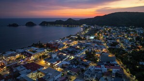 A coastal town at dusk with a long pier extending into the sea, surrounded by residential buildings and a hilly backdrop. - Hotel Tepalcates (San Patricio Melaque)