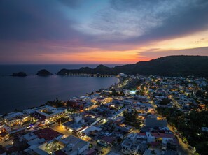 Un pueblo costero al anochecer con un muelle largo que se adentra en el mar, rodeado de edificios residenciales y un paisaje montañoso de fondo.