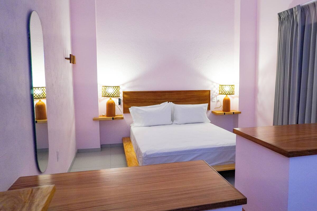 Quarto de hotel com cama de madeira, duas mesas de cabeceira com luminárias, um espelho e janela com cortinas.