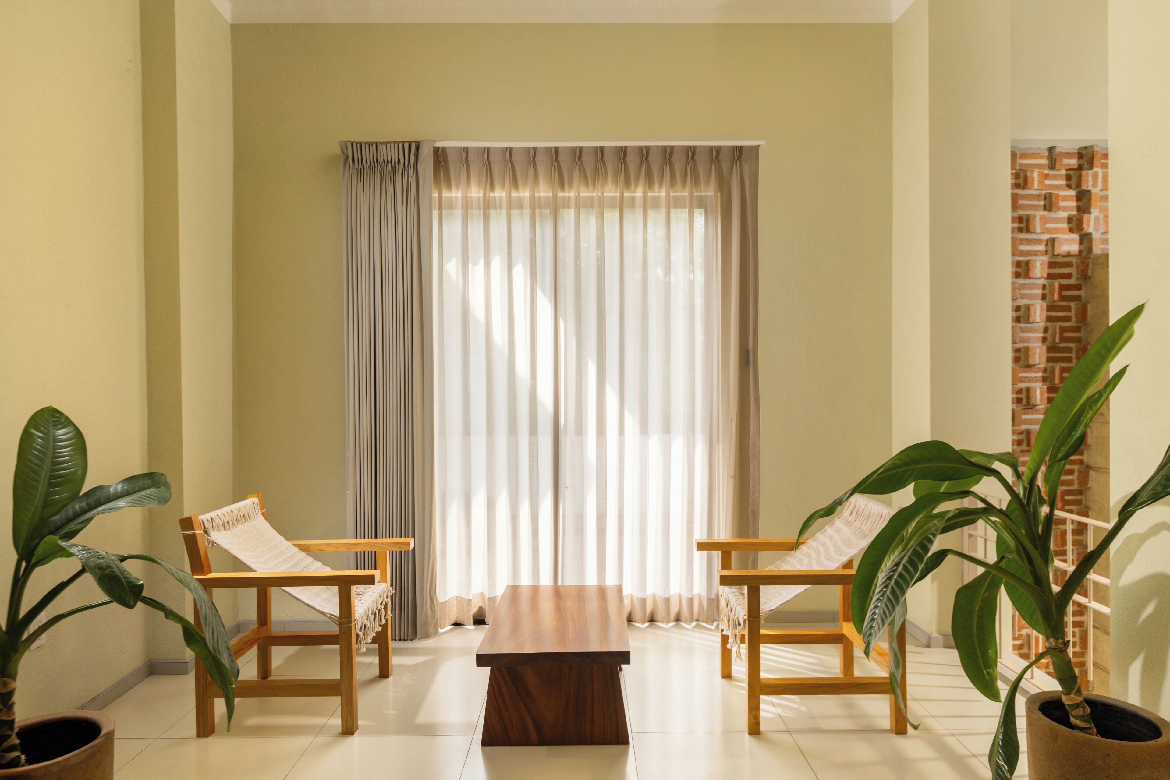 A room with a wooden coffee table, two wooden chairs, a large window with curtains, and potted plants.