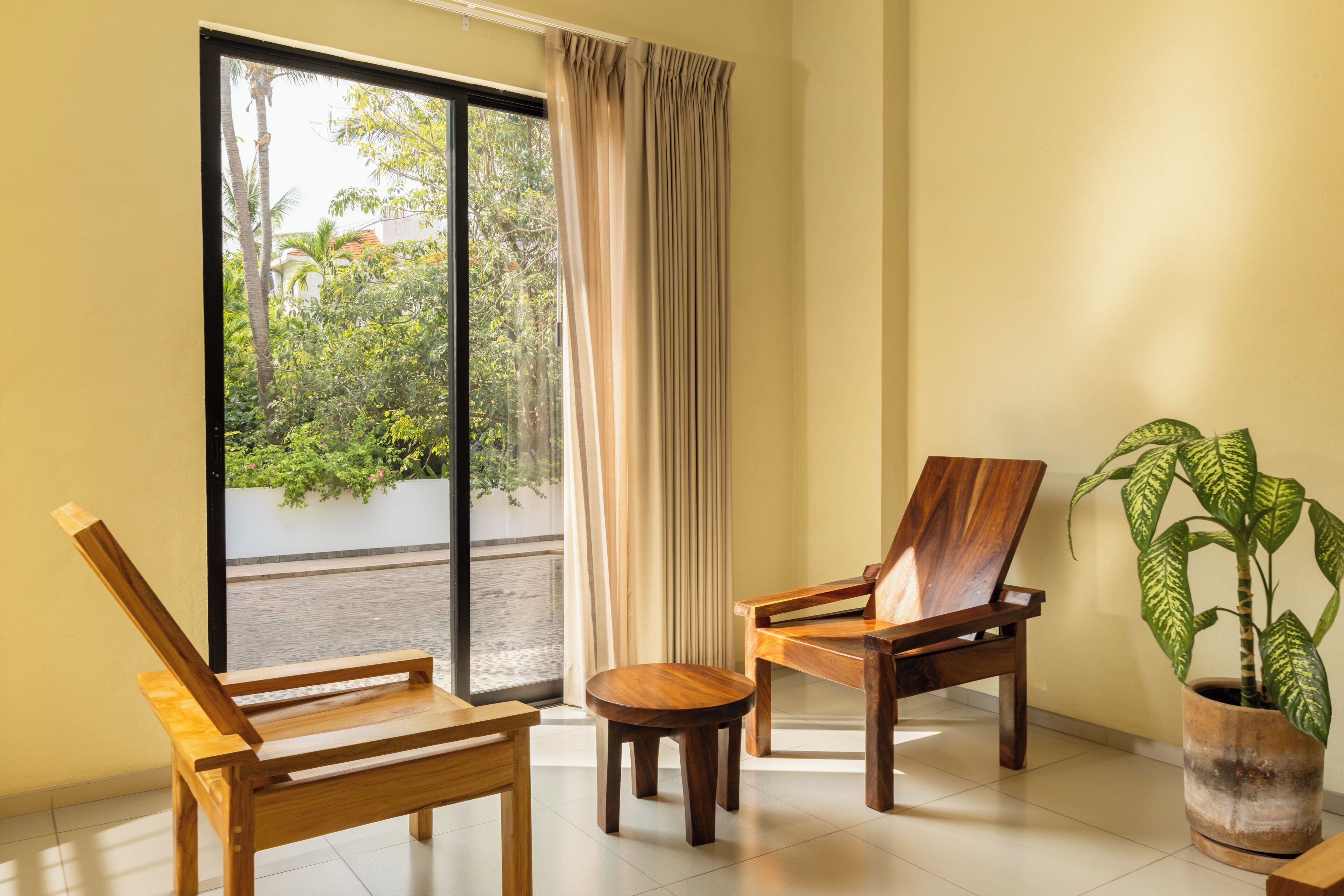 A room with two wooden chairs, a small round table, a potted plant, and a sliding glass door leading to an outdoor area with greenery.