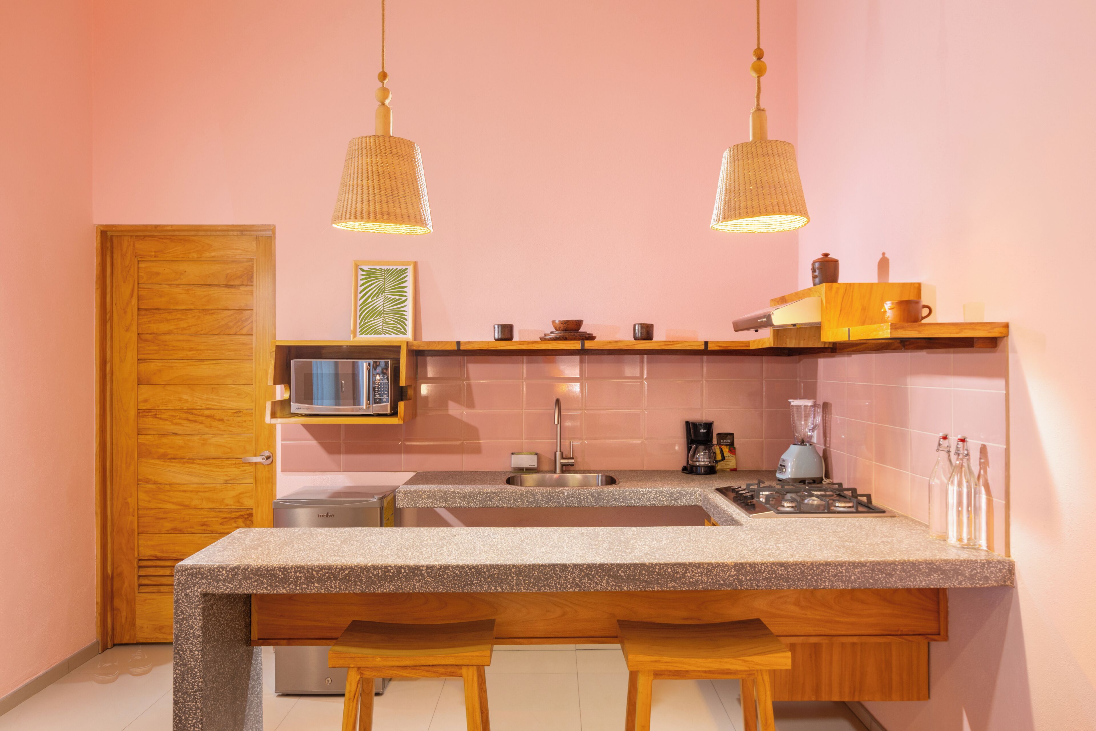 A compact kitchen with a wooden door, a microwave, a sink, and a stove. There are two hanging pendant lights and a small shelf with kitchen items.