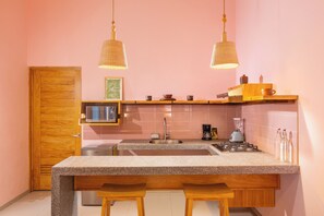 Een compacte keuken met een houten deur, een magnetron, een spoelbak en een kookplaat. Er zijn twee hangende hanglampen en een klein schap met keukengerei.