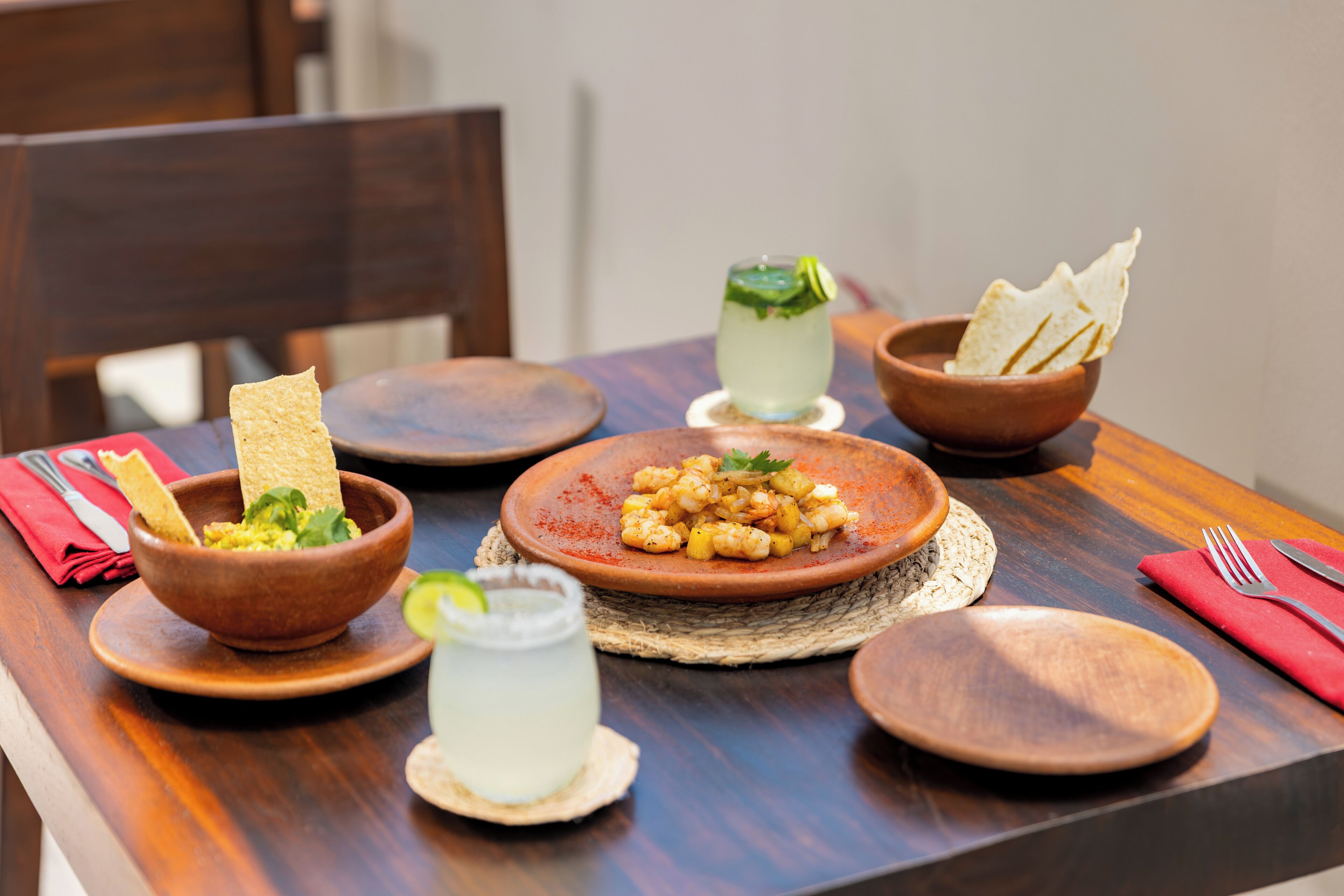 A wooden table with a plate of shrimp, a bowl of guacamole, a glass of limeade, and a bowl of tortilla chips.