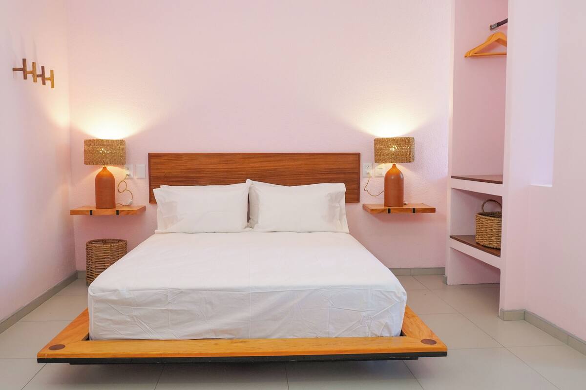 Uma cama com estrutura de madeira e roupa de cama branca, com duas luminárias de parede e cúpulas de tecido trançado.