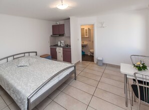 A bedroom with a bed, a small table with a chair, a kitchenette, and a bathroom visible through an open door. - Sunny Elegant Crikvenica Apartments (Crikvenica)