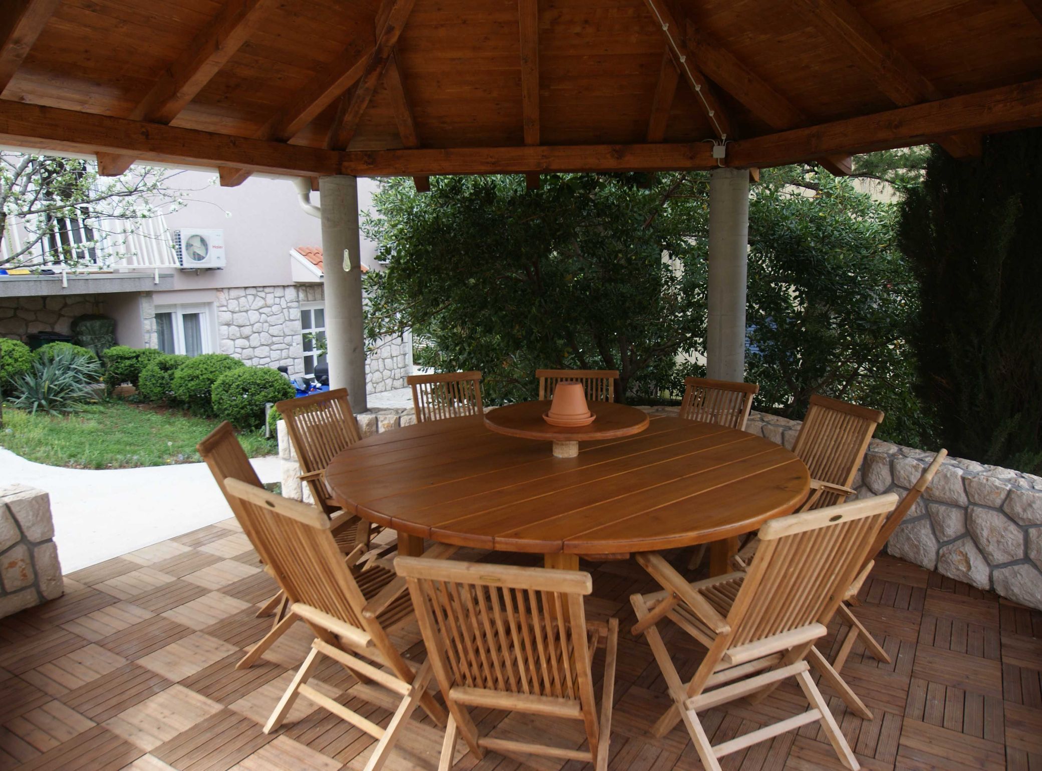 A wooden table and chairs under a covered patio with a stone wall and greenery in the background.