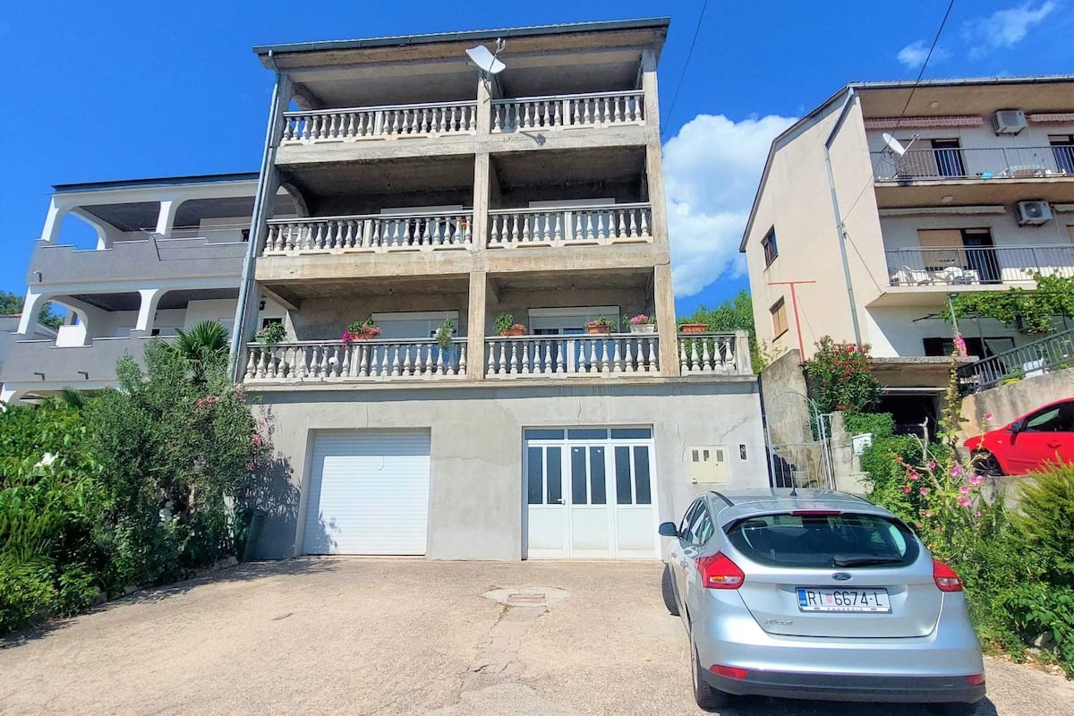 A multi-story residential building with a central entrance, balconies with railings, and a parked car in front.
