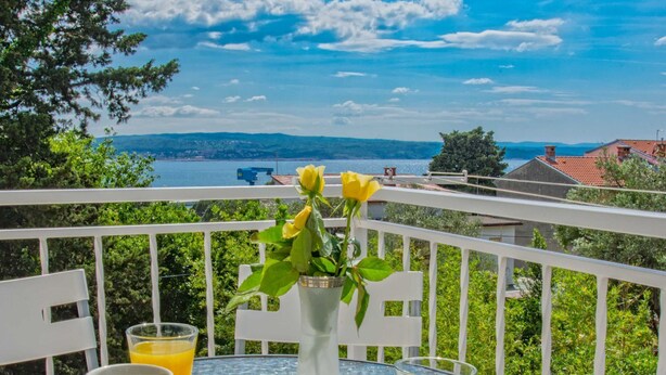 Un balcon avec une table dressée pour deux, deux tasses de café et deux verres de jus d’orange. Un vase avec une fleur jaune est posé sur la table. Le balcon offre une vue panoramique sur un plan d’eau et des collines lointaines.