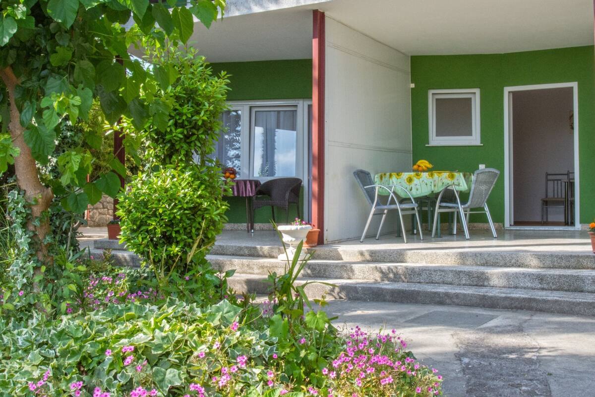 A green building with a covered outdoor seating area, steps leading up to a door, and a garden with pink flowers.