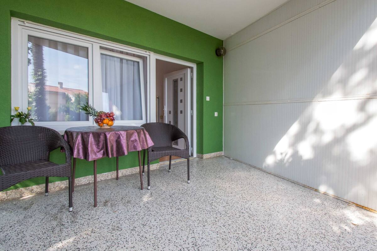 A room with a green wall, a table with a purple tablecloth, two wicker chairs, and a window with a view of trees and buildings.