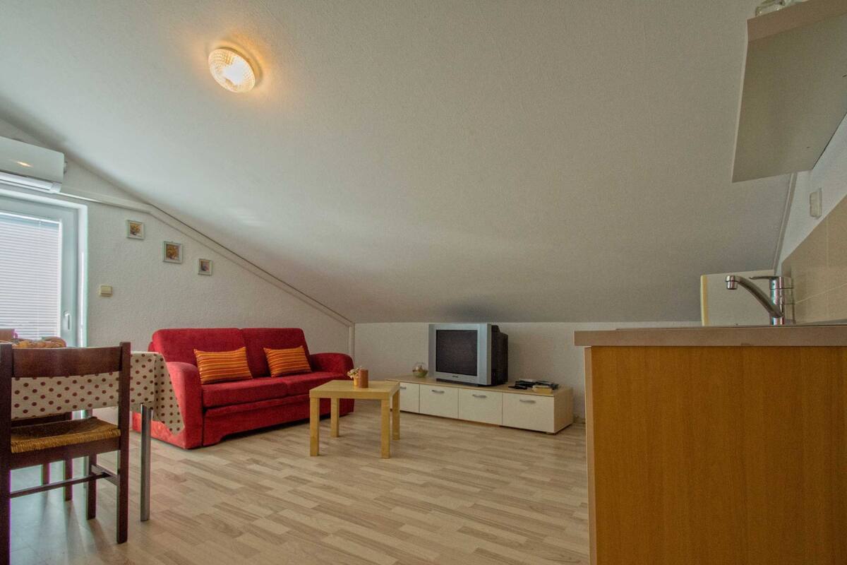 A compact living space with a red sofa, a wooden dining table, a television on a cabinet, and a kitchen area with a sink and countertop.