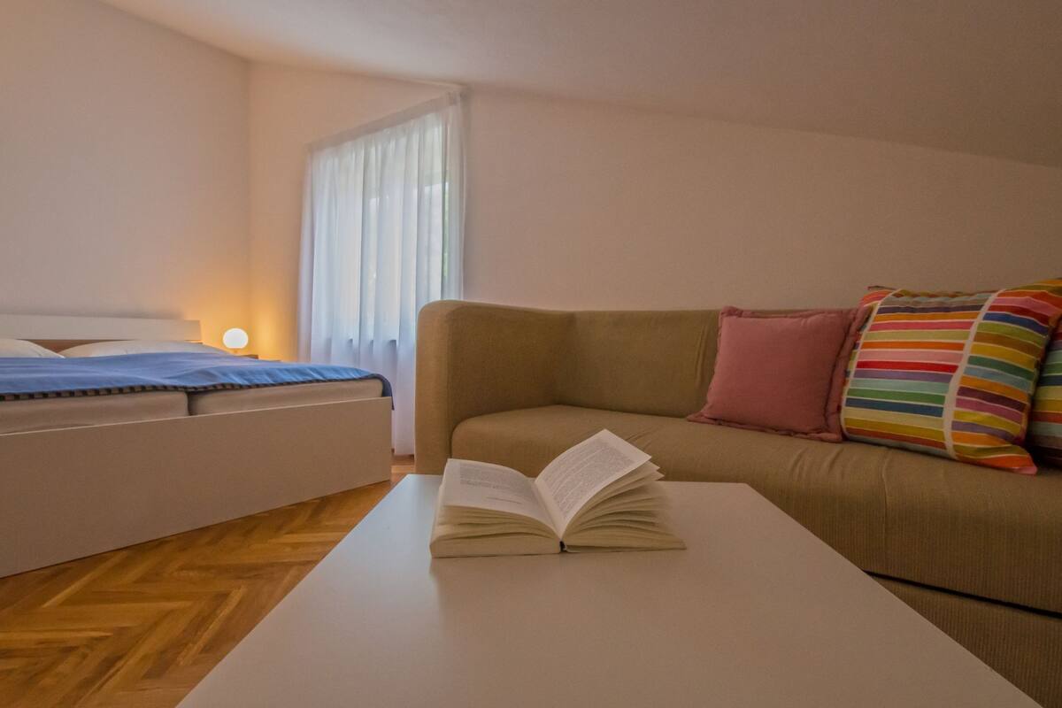 A room with a bed, a sofa, a coffee table with an open book, and a window with curtains.