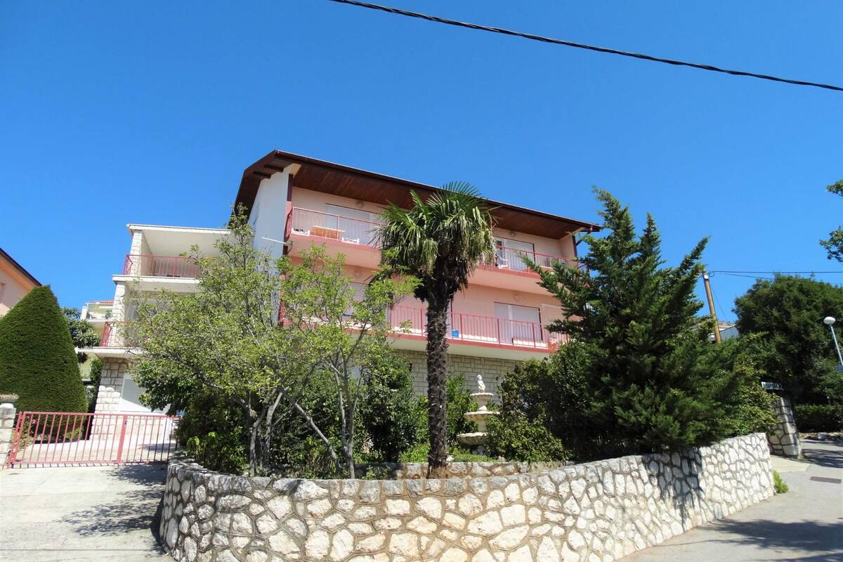 A multi-story building with balconies, surrounded by a stone wall and greenery.