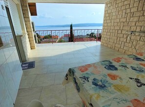 A balcony with a tiled floor, a bed with a floral bedspread, and a view of a body of water and distant hills. - Sleek and Serene Rooms Crikvenica (Crikvenica)