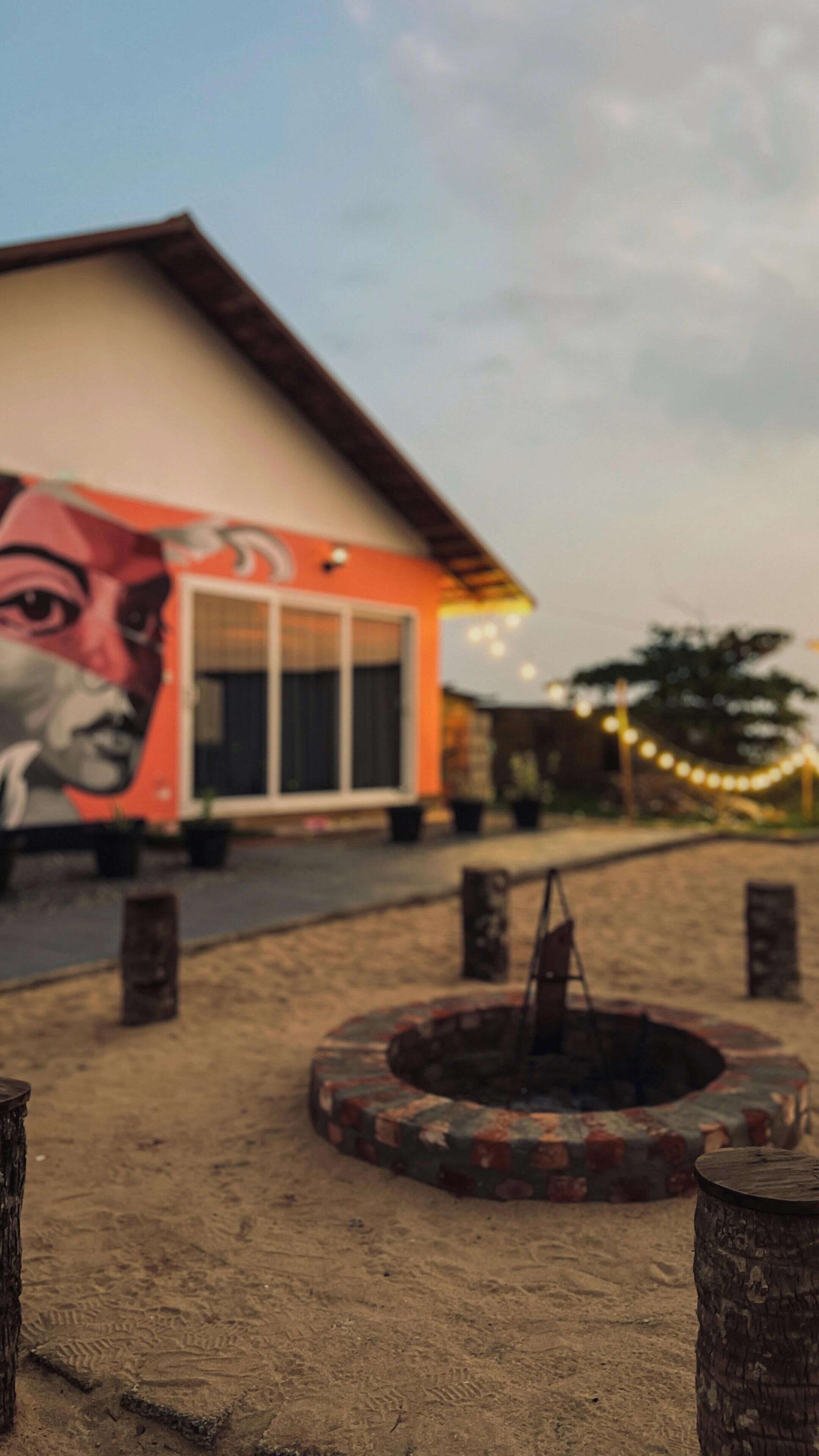 A building with a mural on the side, a fire pit in the foreground, and string lights in the background.