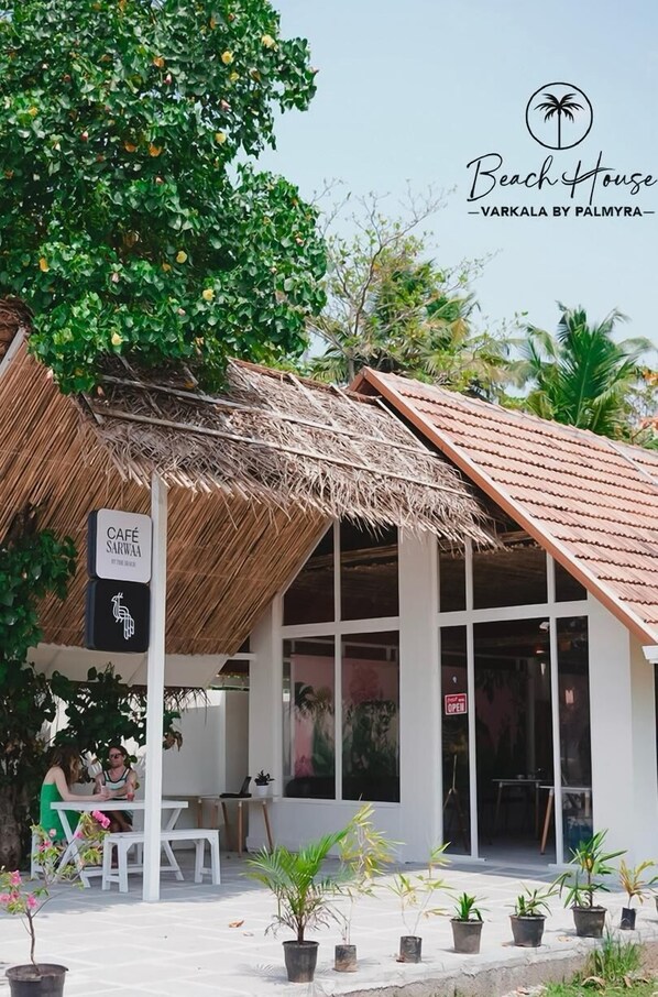 Une maison de plage avec un toit de chaume, une enseigne de café et des plantes en pot.