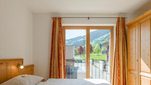 Ein Schlafzimmer mit zwei Betten, einem Holzkopfteil, einem Balkon mit Blick auf einen Berg und ein Gebäude sowie Vorhängen.