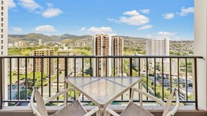 A balcony with a glass table and two chairs, overlooking a cityscape with high-rise buildings and a mountain range.