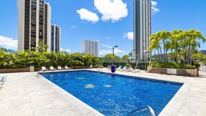 A swimming pool with lounge chairs, surrounded by palm trees and tall buildings.