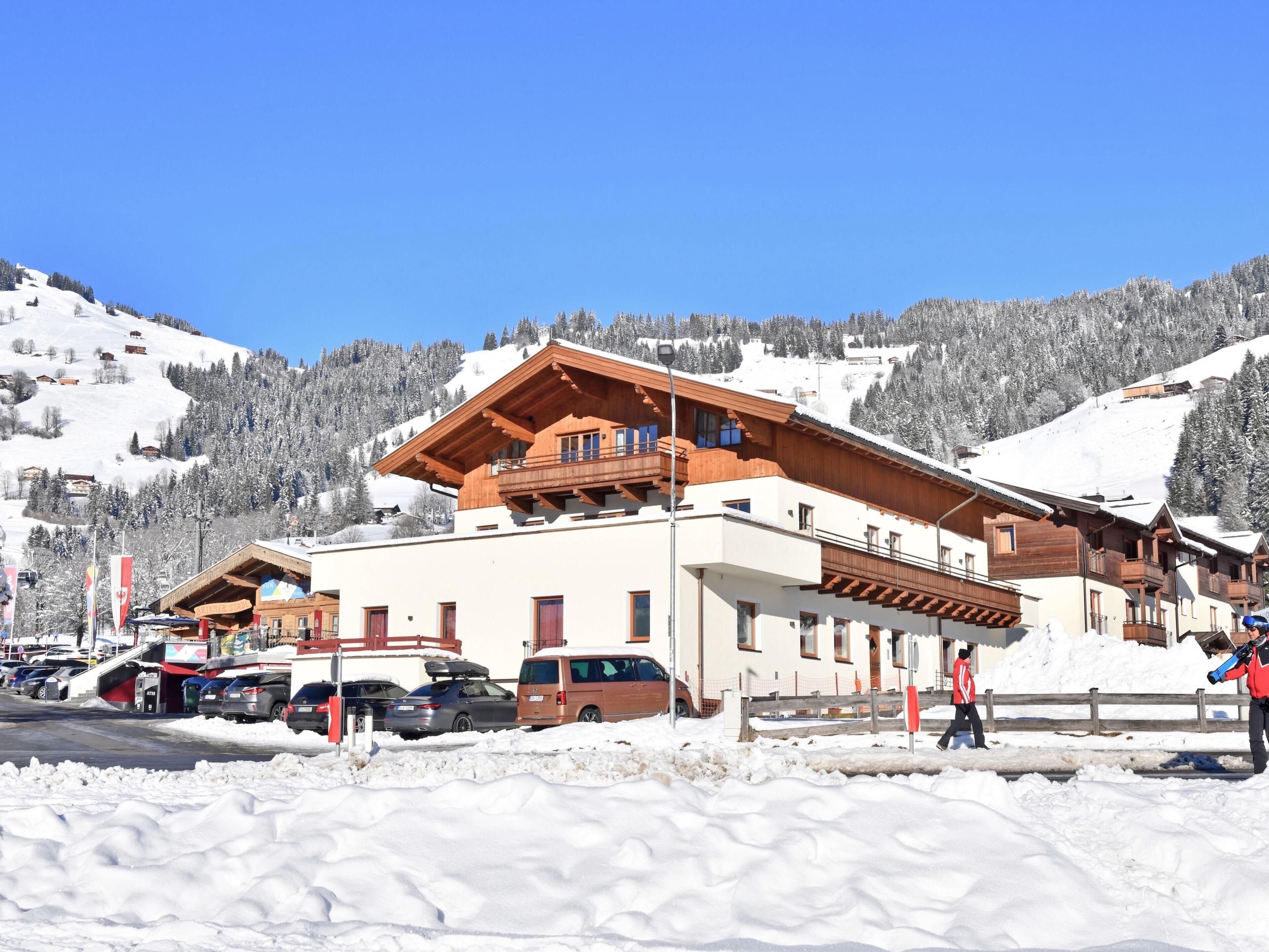 A chalet-style building with wooden balconies, a ski slope in the background, and a parking area with cars.