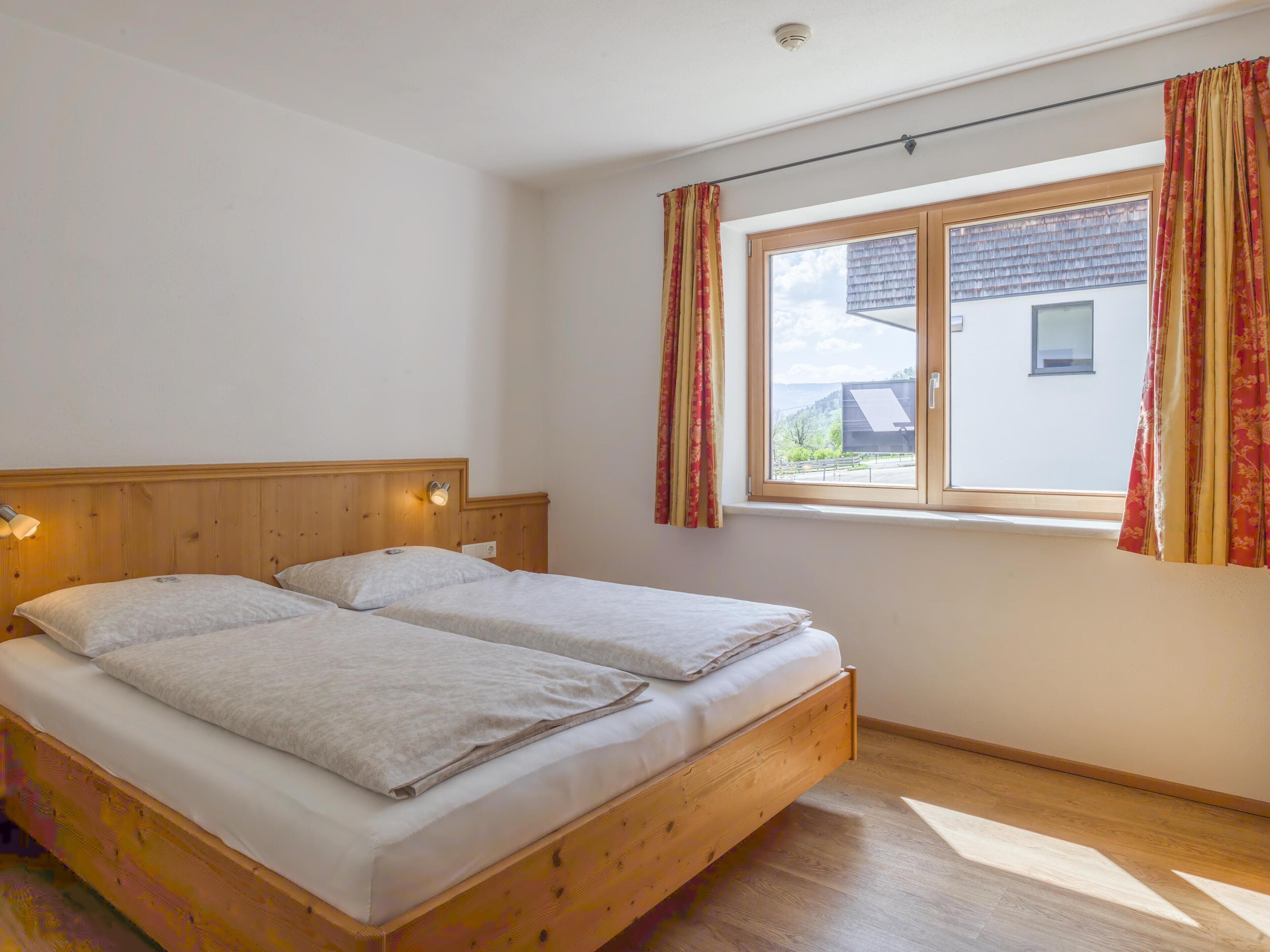 Un lit double avec une tête de lit en bois et des lampes de chevet, une grande fenêtre avec des rideaux et une vue sur un bâtiment à l’extérieur.