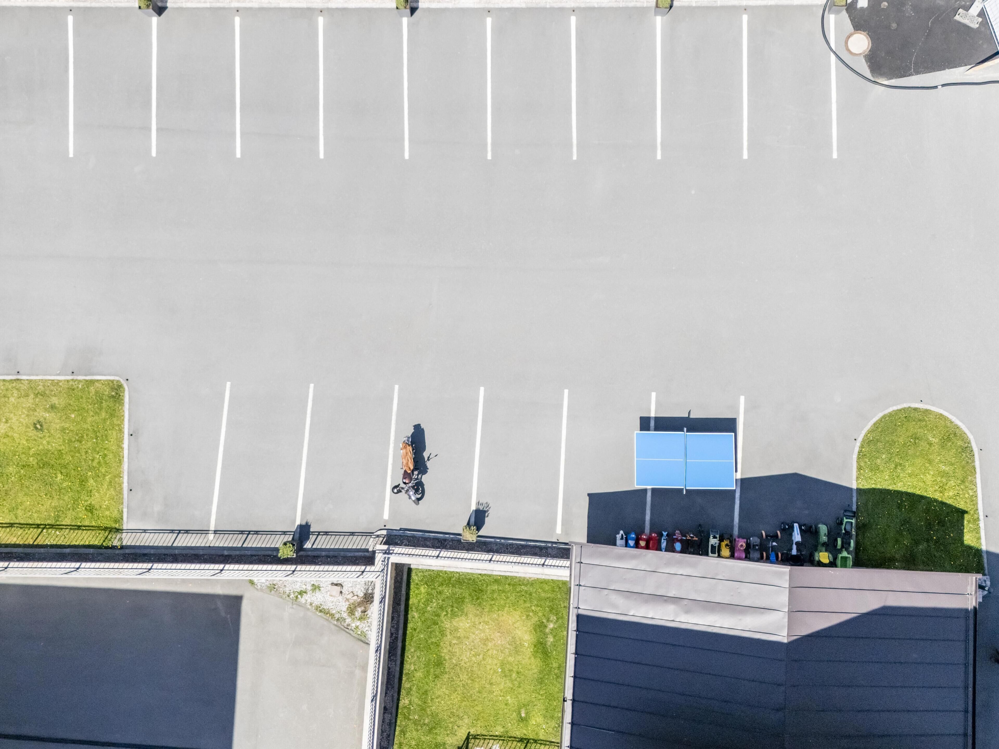 Luftaufnahme eines Parkplatzes mit markierten Stellplätzen, einem kleinen Gebäude mit blauer Tür und einer Rasenfläche.