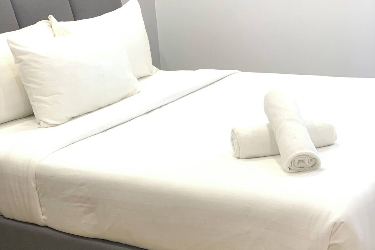 A neatly made bed with white linens and two rolled towels, a grey upholstered headboard, and a light-colored wooden floor.