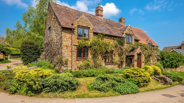 A stone house with a sloped roof, multiple chimneys, and a well-maintained garden with various shrubs and flowers. - Tom Callows Gorgeous Cottage Oxfordshire (Banbury)