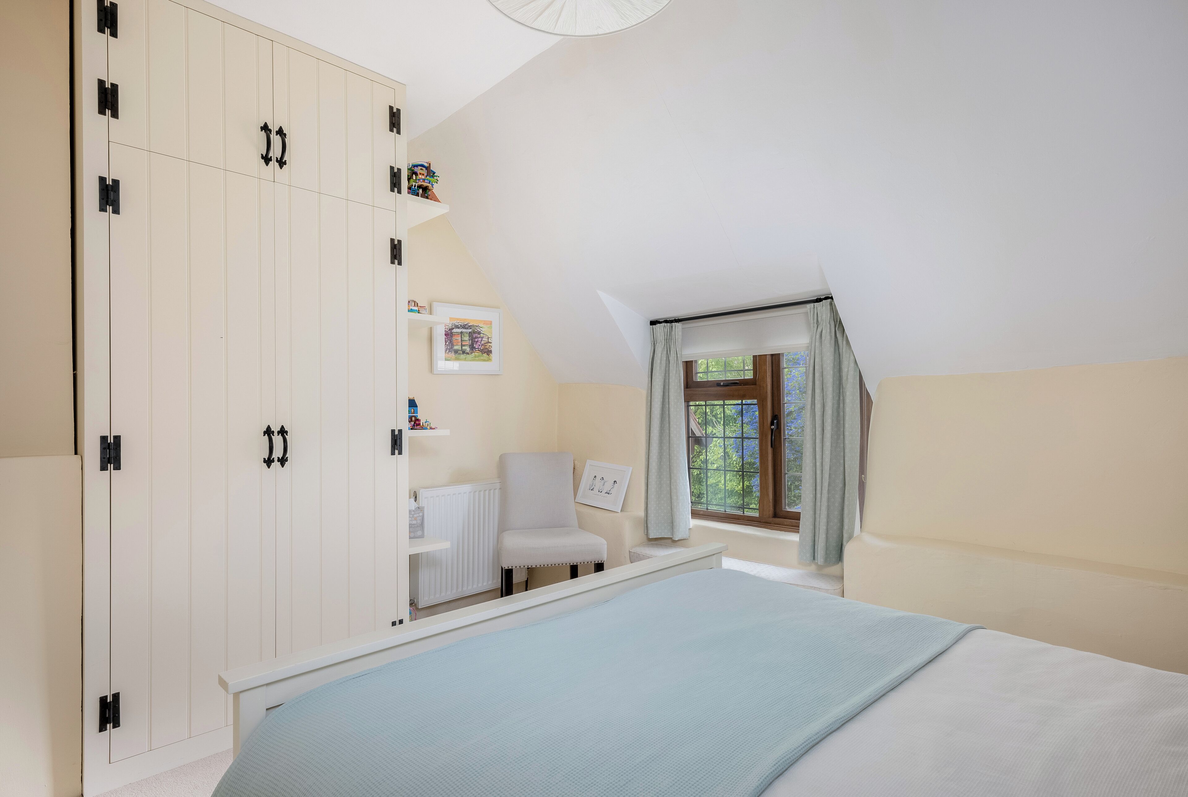 Ein Schlafzimmer mit Bett, Kleiderschrank, Stuhl, einem Fenster mit Vorhängen und einer Heizung.